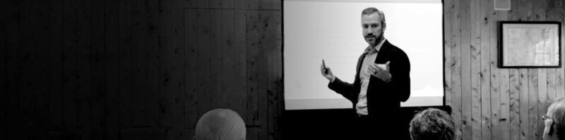 Man presenting to an audience in a wood-paneled conference room